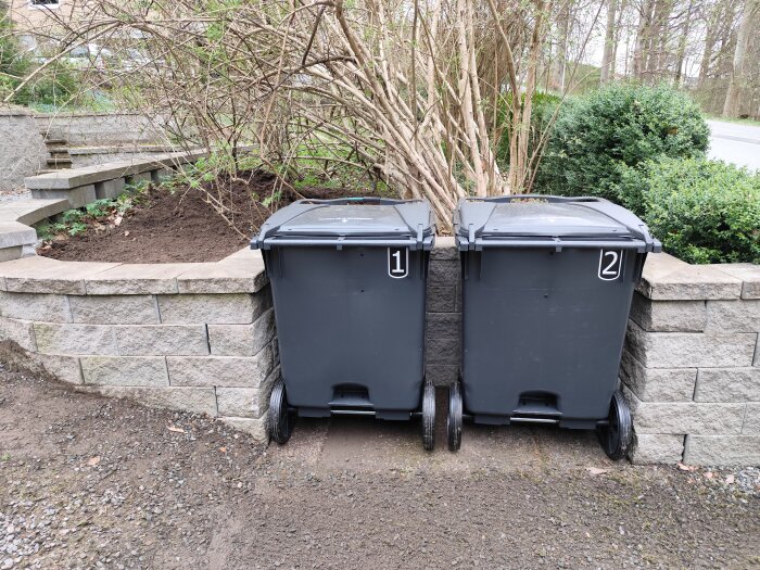 Två gråa soptunnor med nummer 1 och 2 står vid en mur, omgivna av buskar och jord.