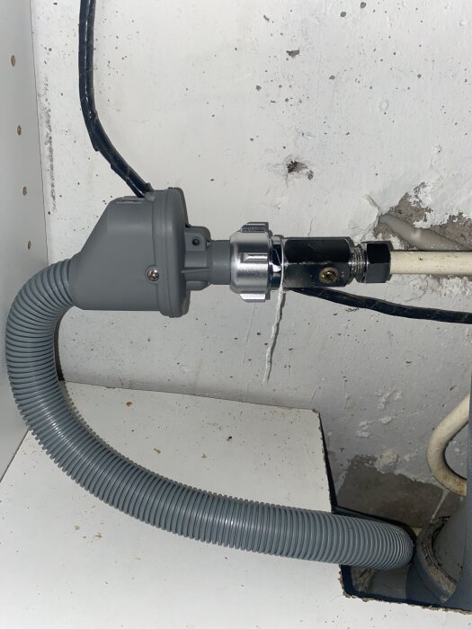 Grå slang ansluten till en aquastop-ventil i köksmiljö, del av diskmaskinens vatteninkoppling, mot vit vägg med synliga rör.