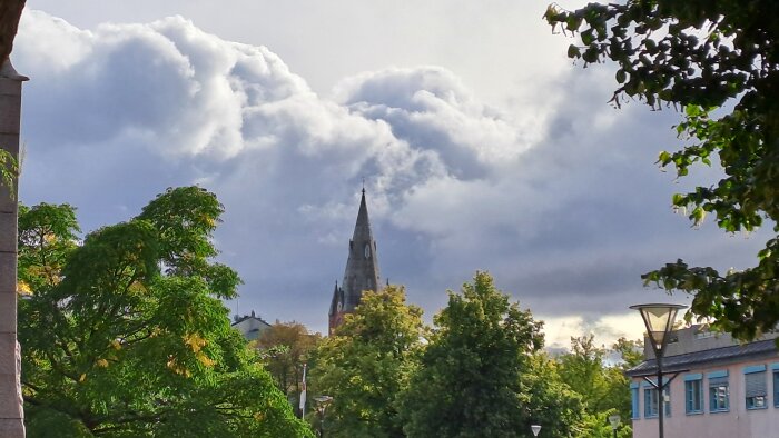 Kyrktorn bakom träd och byggnader under en dramatisk, molnig himmel.
