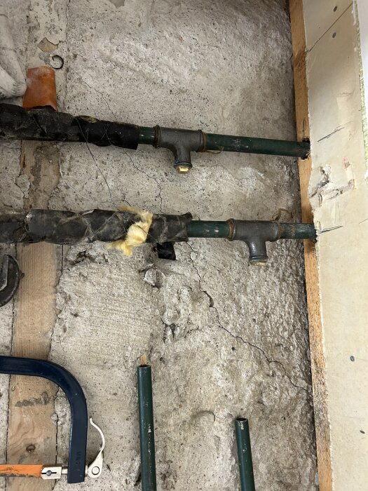 Två gröna rör med mässingsproppar i en vägg av betong. Rören är delvis täckta av svart tejp och omgivna av träreglar.