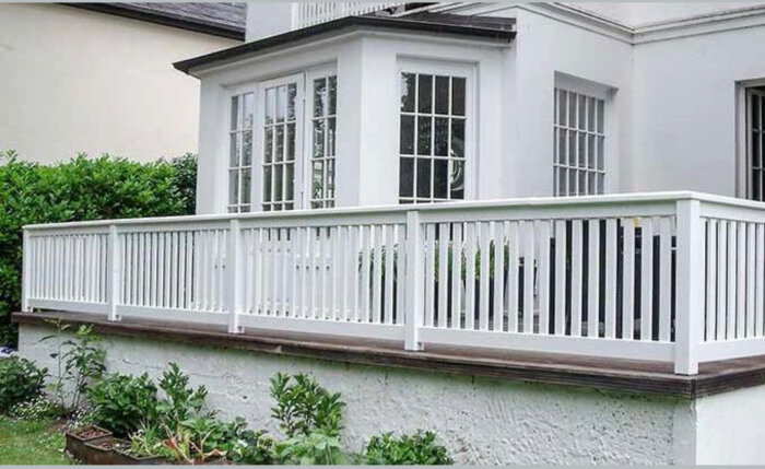 Vit utomhusveranda med räcke och överliggare, fästad vid ett hus med stora fönster och omgiven av gröna buskar.