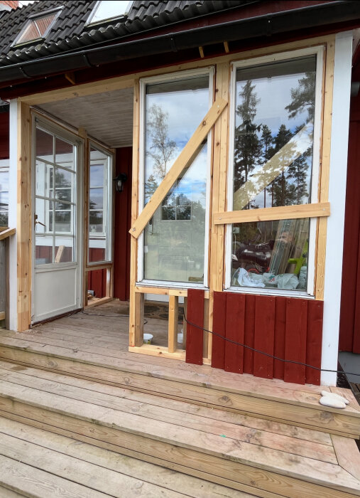 Röd träfasad med träkarmar och tillfälliga fönsterramar där en pardörr planeras. Trätrall framför ingången.