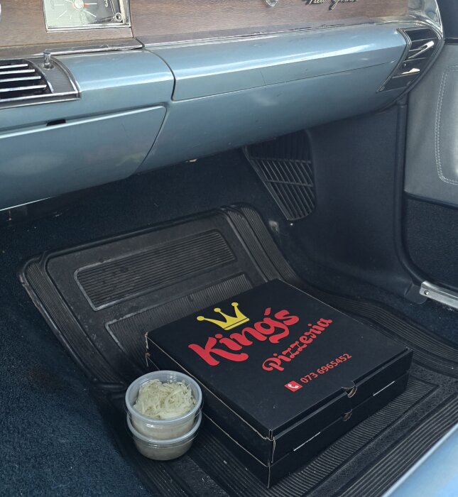 Pizza i kartong från "King's Pizzeria" och två burkar med sås på bilens golvmatta i framsätet.