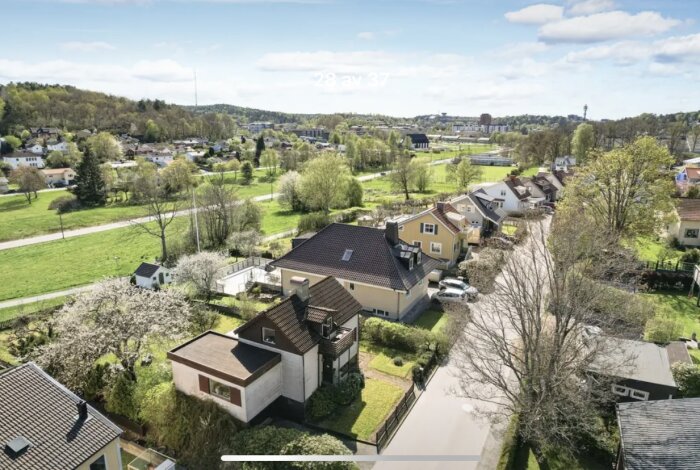 Flygfoto över ett bostadsområde med flera hus, gröna trädgårdar och fält i bakgrunden. Fokus på ett hus med tillbyggnad och garage.