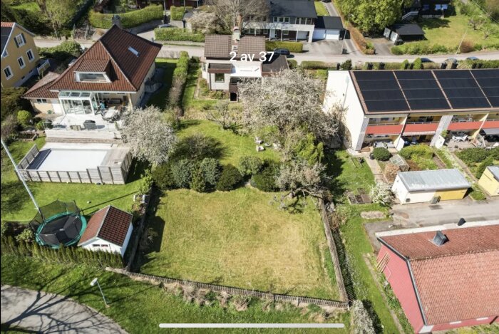 Flygbild över hus med tillbyggnad nära tomtgränsen, omgiven av trädgård och angränsande fastigheter.