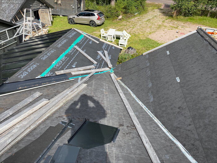 Tak med två ränndalar som möts vid nedre kant, omgivna av byggmaterial och en gårdsplan med en bil och trädgårdsmöbler i bakgrunden.