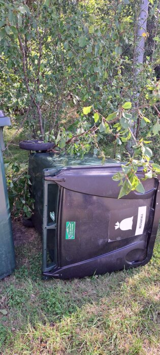 Omkullvält sopkärl med etikett för restavfall, omgivet av gräs och buskar.