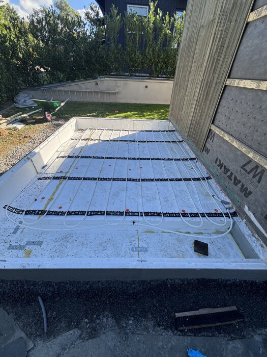 Golvvärmeinstallation på betongplatta utomhus, med slingor synliga. Plattan omringas av växter och byggmaterial.
