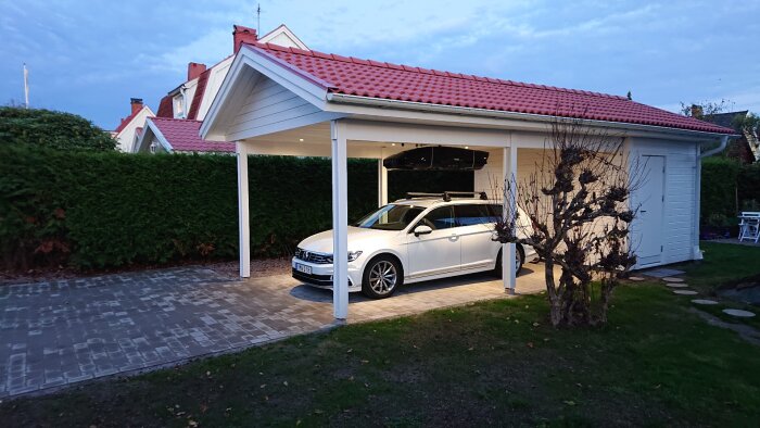 Carport för en bil med röda takpannor, vita väggar och takstolar. Förråd bakom carporten, bil och träd synliga bredvid byggnaden.