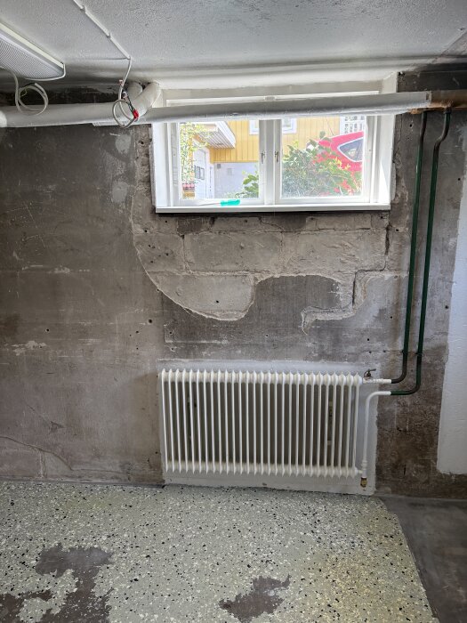 Källarvägg med betonghålsten, synlig spricka under fönster och avskalad puts. Element och rör vid väggen.