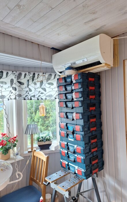En stapel av LBOXX verktygslådor placerade på en stege, som används som ställning för att måla bakom en väggmonterad luftvärmepump.