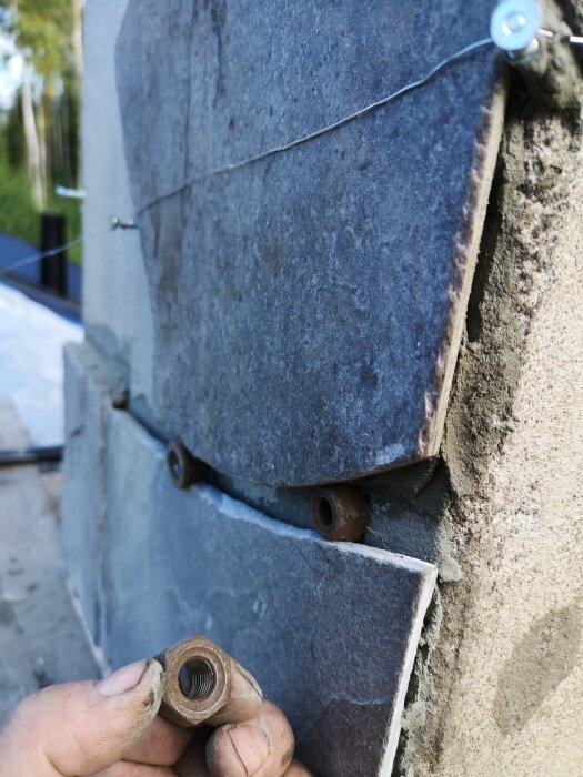 Närbild av en hand som håller i en metallmutter framför en vägg med monterade plattor och infogade metallskruvar.