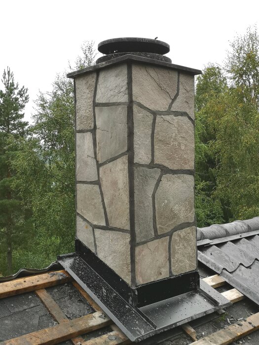 Murad skorsten beklädd med stenplattor på ett tak med svart plåt och takpannor, omgiven av träd i bakgrunden.