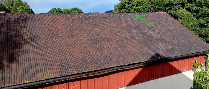 Gammalt plåttak på garage med rost och läckage vid nock, markerat med grön pil. Bakgrunden visar träd och en blå himmel.