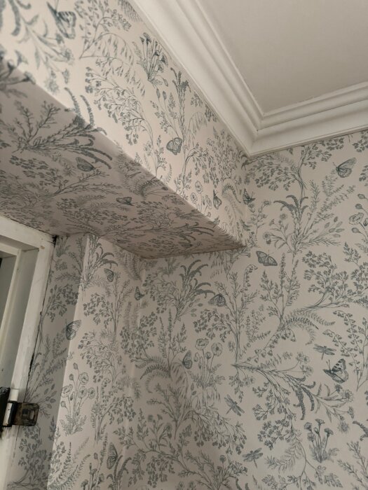 Hörn av ett rum med blommig tapet och vitt taklistverk. Tapeten har mönster av växter och fjärilar.