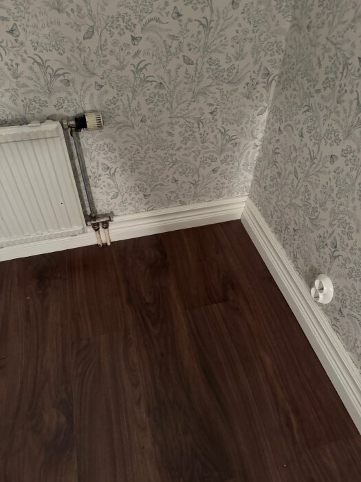 Hörnet av ett rum med mönstrad tapet, mörkt laminatgolv, vitt element och sockel i hörnet. En del av en vit strömbrytare syns på väggen.
