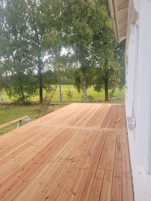 Nybyggd altandäck med träplankor bredvid ett vitt hus, omgiven av grönskande träd med en stege och gräs i bakgrunden.