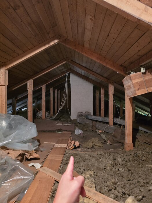 Loftutrymme i hus från 1960-talet med trästommar och takbjälkar, handen pekar mot takstol. Golvet är täckt av byggmaterial och plast.