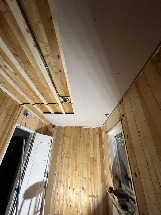 Halvfärdigt gipstak i barnrum med träväggar, en vit dörr och ett fönster.