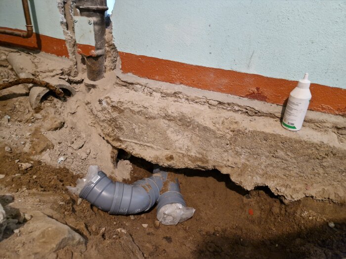 Grävt hål i mark med synliga rör och sönderfallna kanter, flaska på marken.