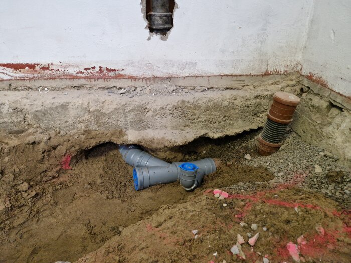 Grävning vid husgrund med exponerade rör och en delvis ihoprasad sektion. Rördelar syns i marken under byggnadens grund.