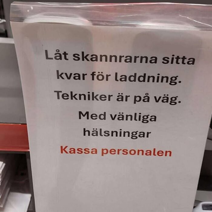 Skylt med meddelande om att skannrar bör sitta kvar för laddning. Med vänlig hälsning från kassapersonalen.