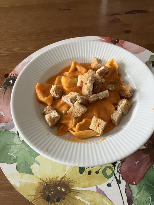 Kelda mustig tomatsoppa med pastakuddar arrabbiata och krutonger i en vit tallrik på ett blommigt underlägg.