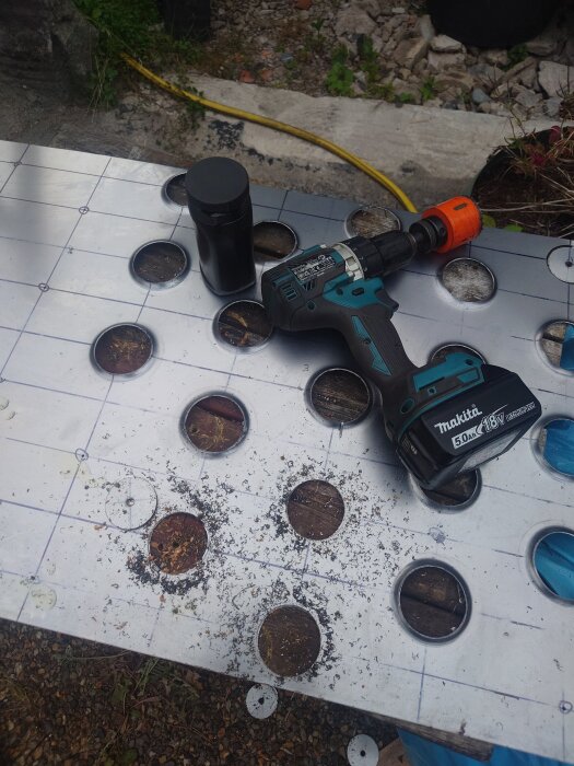 Borrmaskin och borrhuvud på metallplåt med flera borrade hål, omgiven av träspån, utomhus på byggarbetsplats.