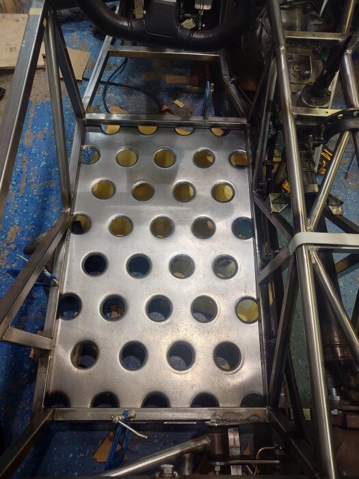 Golvplåt i en bil med 36 runda hål, förberedd för svetsning, omgiven av stålramar och synliga kablar.