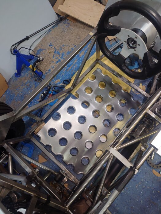 Golvet på förarsidan av ett fordon med hålad stålplåt under konstruktion; synlig ratt och rörstruktur.