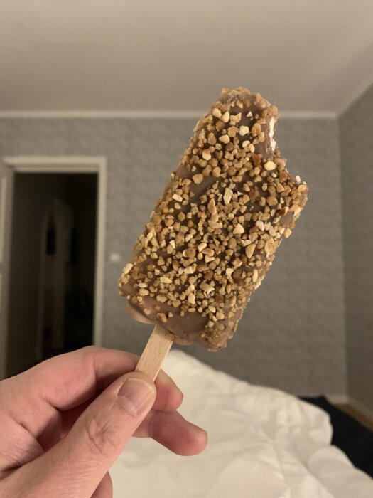 En hand håller en glass på en pinne, täckt med choklad och hackade nötter, i ett rum med en gråmönstrad vägg i bakgrunden.