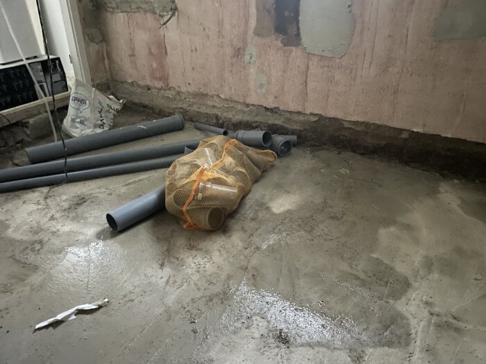Fuktigt källargolv vid väggen, med rör och material på golvet, tydligt mörkare i hörnet där vatten tränger in vid regn.