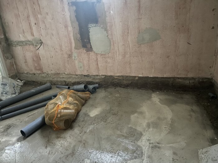 Fuktigt hörn i källare med grå vägg och golv, rör och en orange nätpåse på golvet, visar vatteninträngning mellan vägg och golv.