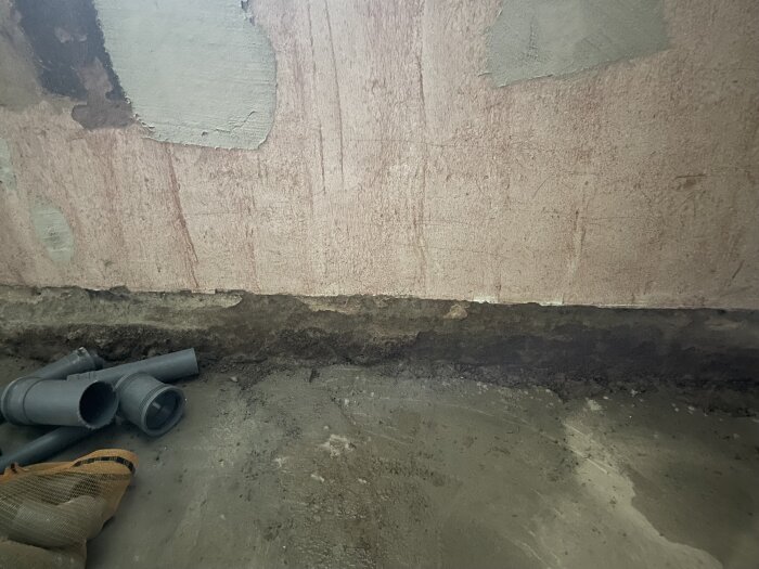 Fuktigt golvhörn i källare med poröst material, mörkare fläck längs väggen, några rördelar i förgrunden, tecken på vatteninträngning.