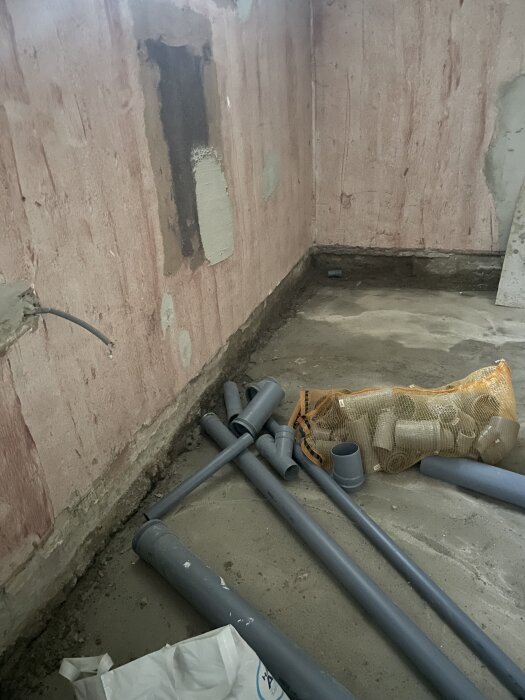 Hörnet av en källare med synlig fukt. Plast- och metallrör ligger på betonggolvet, och väggen visar tecken på avskalning och fläckar.