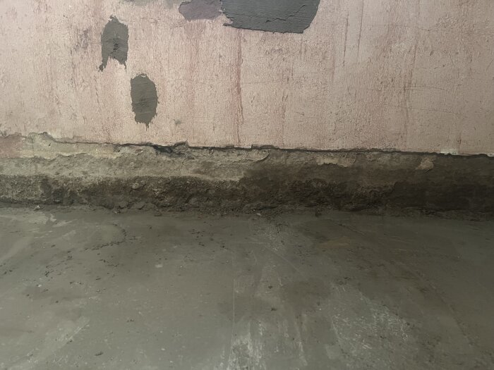 Vägg med fuktinträngning synlig vid skarven mot betonggolv, med flera mörka fläckar och ojämn yta där vatten tränger in vid regn.