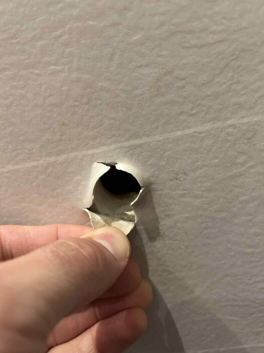 Hål i vägg med trasig tapet runt, uppstått på grund av tidigare eltillförsel. En hand visas som håller i tapetkanten.
