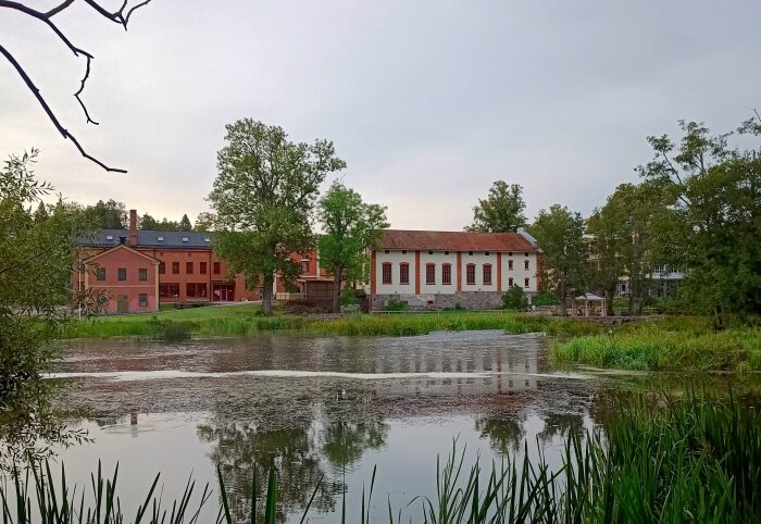 Vy över Rosenfors gamla fabriksbyggnader vid en damm, omgivna av träd och grönska.