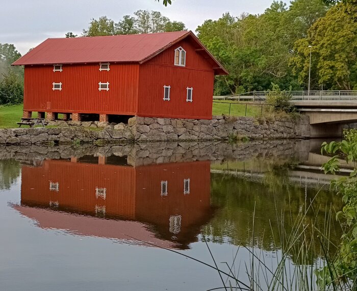 Röd byggnad vid vattendrag med spegelbild i vattnet, omgiven av grönska och stenmur, med bro i bakgrunden.