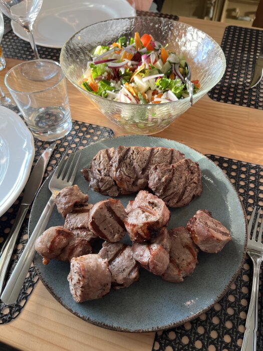Tallrik med grillad fläskfilé och oxfilé, samt en skål med färgglad sallad på ett dukat bord.