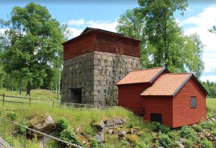 Gammal masugn i Hagelsrum omgiven av grönska, med roströda träbyggnader och stenstruktur, representerar järnbrukets historia.