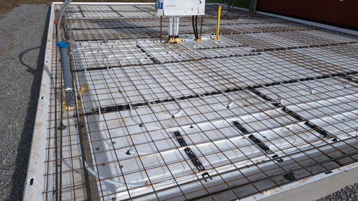 Betongplatta med armeringsjärn och rörinstallationer på en byggarbetsplats, omgiven av grus.