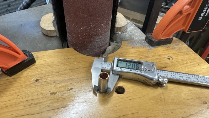 Mätverktyg visar diameter på 12,06 mm bredvid ett metallrör på en träyta vid en slipmaskin.