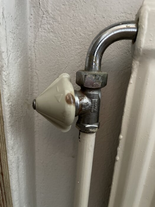 Ventil på en gammal radiator med vit vred och metallanslutning mot väggen, visas i närbild.