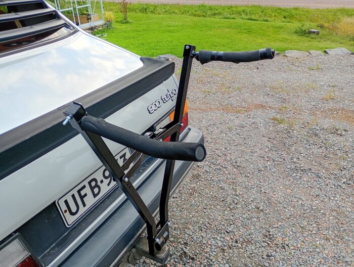 Bilens bakdel med hemmagjord cykelhållare av svartmålat fyrkantsrör och skumgummi, monterad på dragkroken i en grusuppfart.