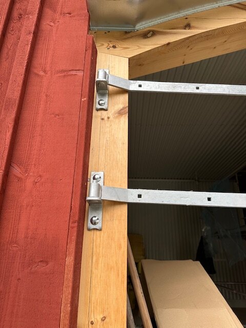 Gångjärn monterade på en träkarm vid en röd träfasad, visar felplacering på grund av dimensionsskillnad i bultarna.