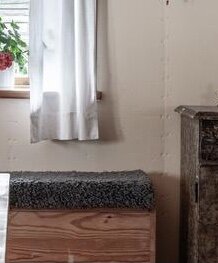 Fäbodens innervägg med ljus panel och fönster med vit gardin, samt antik möbel intill.