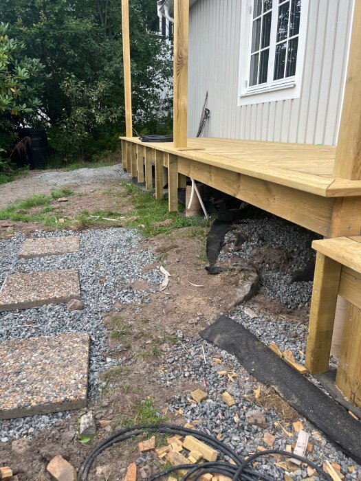 Träterrass under konstruktion med synliga bjälkar, en grusgång med plattor och buskage i bakgrunden.