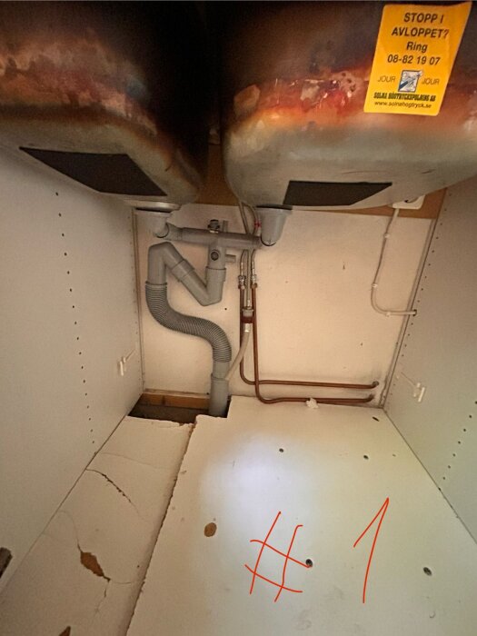 Under en diskbänk med synliga rör och avloppssystem, där gamla bänkdiskmaskinen har stått, med märkning "Stopp i avloppet? Ring 08-82 19 07".