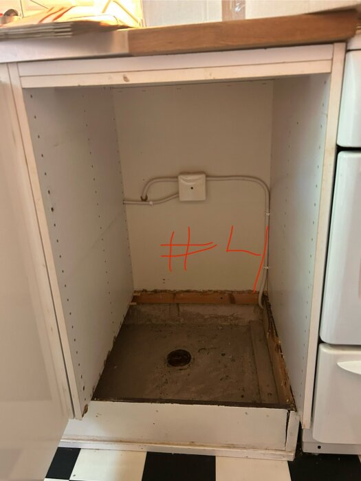 Tomt utrymme i köksskåp för installation av diskmaskin, med ett hål i golvet för avlopp och en synlig elanslutning längs bakväggen.